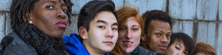 A black woman, asian man, white redhead woman, black man, and asian woman sitting against a grey brick wall