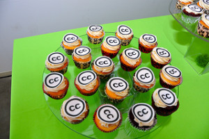 Picture of a circular glass pate on a green table. The plate is covered in multiple cupcakes, each cupcake has a decoration with the letters CC inside a black circle. 
