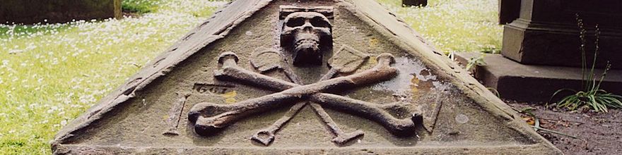 Old coffin shaped stone carved with a skull, crossbones, and spades.