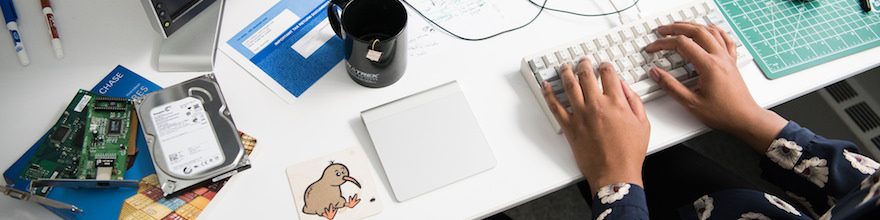 White table with hard drives, a mug of tea, a smart mouse, and keyboard with hands of a black woman typing