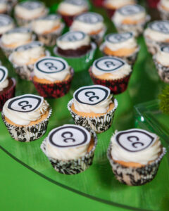 Photograph of cupcakes with white icing and the CC logo on top against a green background. 