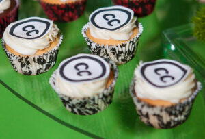 Cropped photograph of cupcakes with white icing and the CC logo on top against a green background.