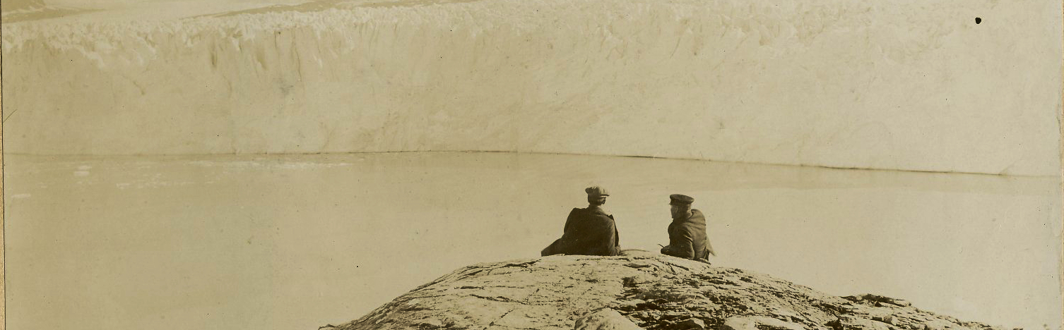 Two men sitting on a rocky cliff looking out across the water at a glacier